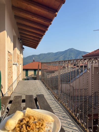 In the city of Orino, Italy, a balcony offers a picturesque view of a mountainous landscape under a clear blue sky. The foreground features a wooden bench and a plate of cereal with banana slices, suggesting a meal enjoyed outdoors. The balcony is enclosed by a decorative wrought-iron railing, overlooking a street lined with buildings featuring terracotta roofs. The sun casts long shadows across the balcony, indicating it is likely late afternoon. The architectural style suggests a Mediterranean setting, with stucco walls and exposed brick accents on the building immediately adjacent to the balcony.