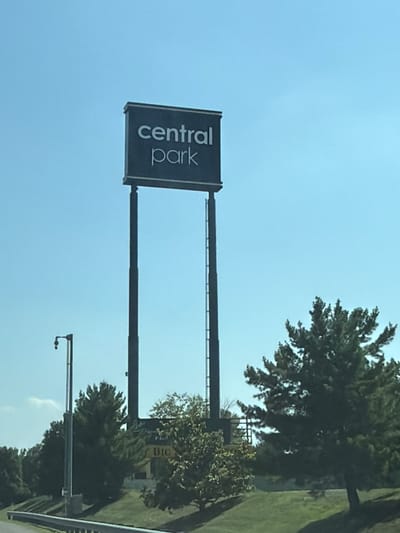 An outdoor, eye-level, medium shot captures a tall sign reading "central park" in white block letters against a clear blue sky. The sign is mounted on two dark, slender poles. To the left of the sign is a street lamp. In the middle ground, trees with lush green foliage are visible. Behind the trees, a yellow building with the word "BIG" in black lettering can be seen. The scene is situated along a road with a guardrail in the foreground, suggesting a location in Fredericksburg, Virginia, United States of America.
