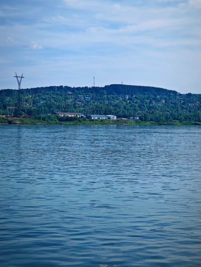 This is a vertical, eye-level, full shot of a wide, blue body of water with a shoreline and a wooded hill in the background. The water is rippled and reflects the cloudy sky. Along the shoreline, there are some buildings, including what appears to be an industrial facility and several houses with blue roofs. A tall, metal power line tower stands on the left side of the frame, and a cell tower is visible on the top of the hill. The hill is covered in dense green trees, with more houses scattered among them. The sky is blue with scattered white clouds. This scene is characteristic of the city of Predivinsk, Russia, situated on the Yenisei River.