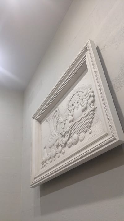 The image shows a close-up, low-angle shot of a white, ornate bas-relief artwork mounted on a textured white wall. The artwork is framed by a decorative border and depicts a still life with a basket overflowing with grapes, a vase, and various fruits and flowers. The lighting suggests an indoor environment, possibly illuminated by a light source from the upper left. The scene is devoid of any people or any indication of a specific location beyond its visual characteristics. There is no information in the image that would allow for the determination of its location in Nikolino, Russia.