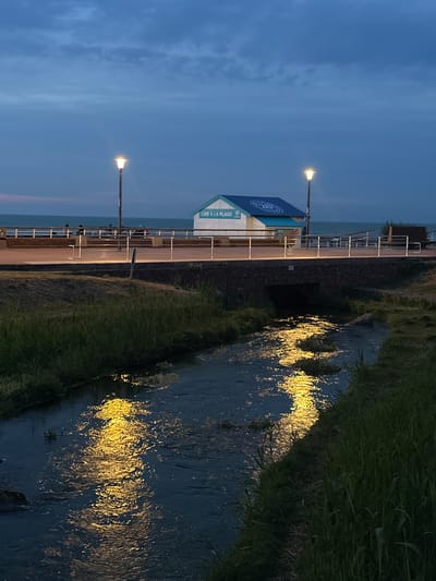 The image depicts a serene evening scene at the mouth of the Veules river as it meets the sea in Veules-les-Roses, France. The sky is a deep twilight blue, with wispy clouds. Two modern streetlights with glowing yellow bulbs illuminate a pedestrian walkway that runs parallel to the coastline. Beyond the walkway, the sea is visible, stretching to the horizon.
In the mid-ground, a small, charming building with a blue roof and white walls stands adjacent to the walkway. It features signage that reads "LIRE À LA PLAGE," suggesting it's a beach library or reading area. Several people are visible on the walkway, seated on benches or strolling, appearing relaxed.
In the foreground, the Veules river flows through a channel lined with lush green grass. The water's surface reflects the warm yellow light from the streetlights, creating shimmering patterns. A small, arched stone bridge crosses the river, with a dark opening visible underneath. The overall atmosphere is peaceful and tranquil, capturing the beauty of the coastal town at dusk.