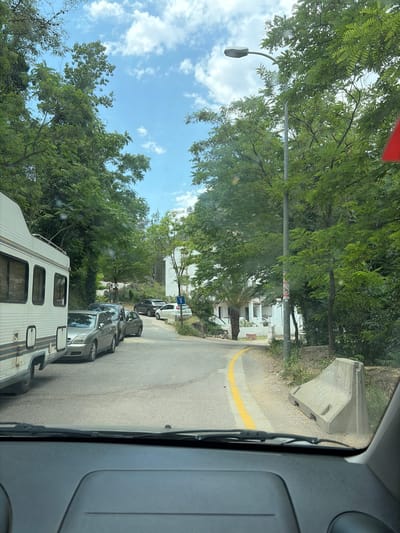 The image depicts a street scene in Montanejos, Spain, taken from inside a vehicle. The vehicle is parked on a narrow road, with other cars parked in front. On the right is a gray concrete barrier. A yellow line marks the edge of the road. Tall green trees line the street, partially obscuring white buildings. A streetlamp is visible on the left. The sky is visible through the trees, showing a mix of blue and white clouds. In the lower portion of the frame is an RV.