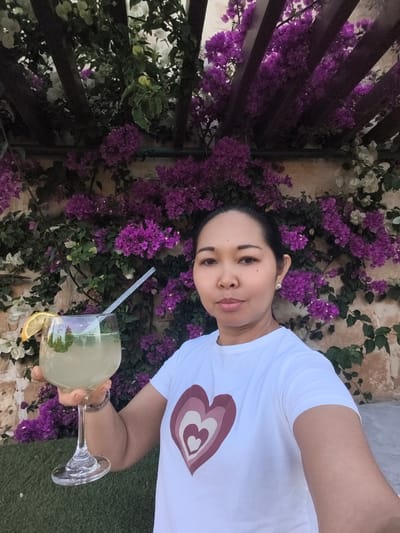 A medium indoor shot depicts a woman standing in front of a wall covered in purple and white bougainvillea. The wall is adorned with wooden trellis. The woman is holding a large glass of what appears to be lemonade, garnished with a lemon slice and a straw. She is wearing a white t-shirt with a heart graphic. Her hair is pulled back, and she is wearing small pearl earrings. The location is San Ġwann, Malta, indicated by the architectural style and the prevalent bougainvillea.