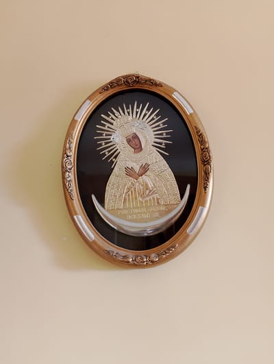 The image shows a framed icon of the Virgin Mary hanging on a light beige wall. The icon is oval-shaped with a gold-colored frame featuring floral motifs. The Virgin Mary is depicted with a halo and a crown, wearing a richly ornamented robe. She is standing on a crescent moon, and her hands are clasped in front of her chest. Below her image, there is text in Polish, which reads "POD TWOJĄ OBRONĘ UCIEKAMY SIĘ". This translates to "Under your protection, we flee". The icon is centrally located on the wall, with ample empty space around it. The lighting appears to be natural, coming from the right side of the frame, casting a slight shadow on the left. The scene suggests a religious or devotional setting, possibly a home or a church.
The image likely originates from Kolno, Poland, as indicated by the Polish text on the icon, which is a common devotional phrase associated with the Black Madonna of Częstochowa, a revered icon in Poland. While the specific location within Kolno cannot be determined from the image alone, such icons are typically found in churches, chapels, or private residences in Polish towns and cities.