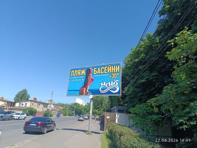 The image shows a street scene in Odesa, Ukraine, on a sunny day. A large blue billboard with yellow lettering and an image of a woman in a pink bikini is prominently displayed. The billboard advertises "ПЛЯЖ БАСЕЙНИ" which translates to "Beach Pools" and mentions "з морською водою +30°" meaning "with seawater +30°". Below this, it lists a phone number "050 338 91 98" and the name "НЕМО ПЛЯЖНИЙ КОМПЛЕКС" which translates to "NEMO Beach Complex".
On the street, several cars are visible. A dark blue sedan with license plate BR0747XX is parked on the right side of the road. Further down the road, other vehicles, including a white hatchback and a grey SUV, are driving or parked. Pedestrians are also present on the sidewalk. The environment includes trees and buildings lining the street, with power lines crossing overhead. A timestamp in the bottom right corner reads "22.06.2026 14:49", indicating the date and time the photograph was taken.