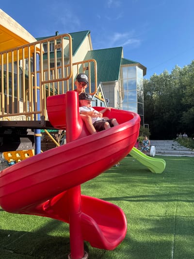 A medium shot captures two young boys on a playground slide in the city of Danilovka (Altai Krai), Russia. The boy in front, wearing a white t-shirt and dark shorts, is seated on the bright red, spiral slide, looking down. Behind him, an older boy in a red t-shirt and a black baseball cap, also looking forward, has his arm around the younger boy. The playground equipment, a mix of red and yellow plastic and yellow metal railings, dominates the foreground. In the background, a building with a green metal roof and large windows is visible, surrounded by trees. The sky is clear and blue, with some wispy clouds. The ground is covered in green artificial turf.