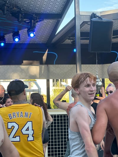 The image shows a group of people gathered outdoors, possibly at an event or concert. A young person with light-colored hair and a gray tank top is in the foreground, smiling and looking to the right. To the left, a person wearing a yellow Kobe Bryant #24 jersey is visible from the back. In the background, there is a stage area with blue lights and a patterned ceiling. The setting appears to be a public gathering space with an open-air roof structure. There are no indications that this image is located in Borgonato, Italy.