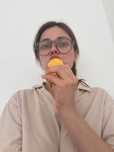 A woman with dark hair and wearing round glasses holds a yellow object to her mouth. She is wearing a light beige collared shirt. The background is a plain white wall. The location is unknown, as the image provides no information about Kőröshegy, Hungary.