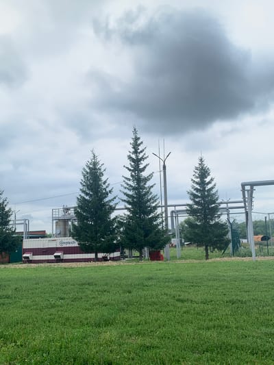 This image shows an outdoor industrial site in Rakashevo, Russia. In the foreground, a lush green field of grass dominates the lower half of the frame. Behind the field, a series of industrial structures are visible. These include a large, rectangular container unit with maroon and white stripes, bearing the text "Comptech." To the right of this unit, several tall evergreen trees are positioned in a line, with their branches reaching towards the sky. Behind the trees, a complex network of pipes and scaffolding is apparent. The background features a cluster of smaller buildings and more greenery, suggesting a rural or semi-industrial area. The sky above is overcast with heavy, dark grey clouds, indicating a cloudy or potentially stormy day. There are no people visible in the image.