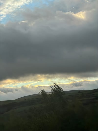 This image captures a dramatic natural landscape, likely in the region of Blajievo, Bulgaria, dominated by an expressive sky at either sunrise or sunset.
The scene is composed of a contrasting sky and a rolling, hilly terrain. The upper portion of the sky is covered by extensive, dense dark grey clouds, suggesting heavy cloud cover or an approaching weather front. Below these dark clouds, a spectacular, horizontal band of intense golden light cuts across the mid-ground, illuminating the edges of the clouds with vibrant hues of yellow and orange. This brilliant light appears to be breaking through or reflecting off the clouds, creating a striking visual division. To the far left, patches of lighter, whiter clouds and glimpses of a pale blue sky are visible, hinting at clearer conditions beyond the main cloud mass.
Below the luminous sky band, the landscape consists of dark green, undulating hills or mountains, partially silhouetted against the brighter horizon. The terrain appears rural and natural, with no visible structures or human activity. In the immediate foreground on the right side, the dark silhouette of a tree branch with foliage extends into the frame, adding depth to the composition.
No people, specific objects (other than natural elements), or text are visible in the image. The dramatic lighting and cloud formations evoke a sense of awe and highlight the powerful beauty of natural weather phenomena, characteristic of either dawn or dusk.