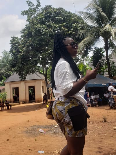 A person stands outdoors in Umundugba, Nigeria, holding a mobile phone. The individual wears a white t-shirt, patterned shorts, and sunglasses. The environment includes a building with window openings, trees including a palm tree, and bare ground littered with plastic bottles. Other individuals are visible in the background.