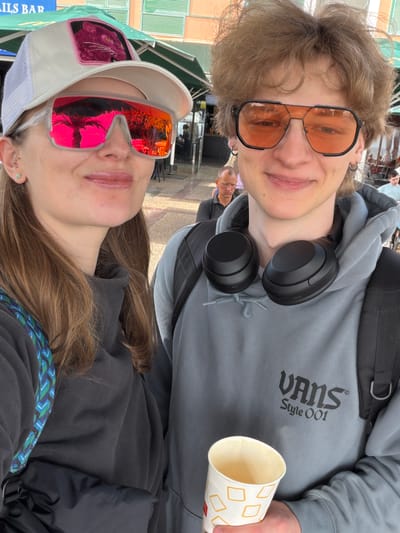 A selfie-style image, rotated 90 degrees counter-clockwise, features two young individuals smiling at the camera in what appears to be an outdoor public setting, possibly Los Cristianos, Spain.
The person positioned at the top, a male with light brown hair, wears large orange-tinted aviator-style sunglasses that reflect parts of the environment. He is dressed in a grey hoodie, which clearly displays the black "VANS" logo with "Style OO1" underneath it on the chest. Black headphones rest around his neck, and a black backpack strap is visible over his right shoulder. He holds a white paper cup patterned with orange squares in his right hand.
Below him, a female with long brown hair also smiles at the camera. She wears striking mirrored pink and orange sunglasses, which show reflections of what appears to be trees or palm fronds. Her head is adorned with a white trucker hat featuring a mesh back and a distinctive pink panther patch on the front. She is wearing a dark grey or black jacket, and a blue and green patterned strap is visible over her shoulder. A small ear piercing is visible on her left ear.
The background is somewhat blurred but shows glimpses of buildings, green structures like awnings or blinds, and other indistinct figures, indicating a bustling public space. A blue sign with partial text "AILS BAR" is visible behind the female subject. The bright lighting suggests it is daytime, and the presence of sunglasses on both individuals implies sunny weather. Both subjects exhibit cheerful expressions.