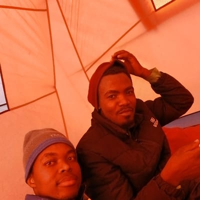 This image depicts two adult males inside a distinctly orange-red tent, located at Gilman's Point, Tanzania. The man on the left, positioned closer to the camera, wears a grey beanie with a blue stripe, a blue jacket, and looks directly forward. He has a dark, short beard and mustache. The man on the right, situated slightly behind and to the right, wears a red beanie, which he is adjusting with his right hand. He has a dark, short beard and is dressed in a dark, possibly black, puffer jacket displaying a light-colored logo on the left chest, with a light-colored shirt collar visible beneath. His left hand is partially visible, holding an unidentified object. Both subjects are looking towards the camera. The tent's interior exhibits visible structural seams and lines against the orange-red fabric. A red fabric object is partially visible behind the man on the right.