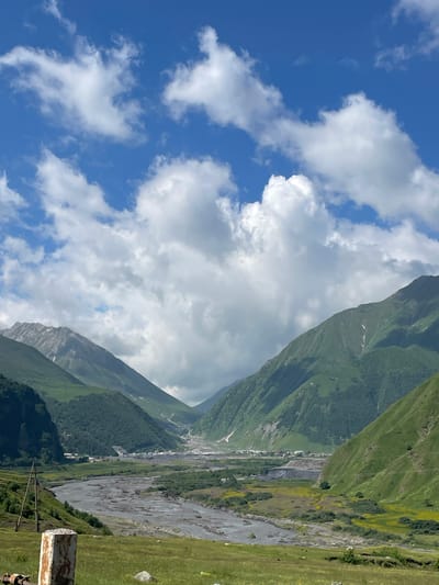 The image depicts a wide, scenic valley in Kanobi, Georgia, under a bright blue sky dotted with fluffy white clouds. Lush green mountains rise steeply on either side of the valley, their slopes covered in dense vegetation. A winding river, appearing somewhat muddy, flows through the center of the valley floor, flanked by areas of grass and scattered yellow wildflowers. In the distance, a small settlement is visible, with buildings clustered together amidst the vast landscape. A rustic wooden utility pole stands tall on the left side of the foreground, and a weathered concrete marker with exposed rebar is positioned in the lower left corner, suggesting a roadside view. The overall scene conveys a sense of natural grandeur and remote tranquility.