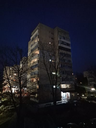 The image displays a nighttime scene in Odorheiu Secuiesc, Romania, featuring a prominent multi-story residential building under a dark blue sky. The central structure is an approximately 10-12 story apartment block constructed of light-colored concrete or stucco. Numerous windows across its facade are illuminated from within, indicating habitation, with a brightly lit main entrance visible on the ground floor. Several bare deciduous trees with visible branch patterns are positioned in the foreground and midground, partially obscuring the main building and a less illuminated, smaller multi-story building to its left. Two park benches are visible near the main building's entrance. The lower right portion of the frame shows dimly perceived vehicles in a parking area, while a terraced landscape with steps is present on the lower left. No individuals or specific activities are discernable within the captured scene.