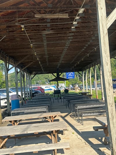 The image shows an outdoor covered pavilion at an unspecified location, possibly a park or recreational area. The pavilion has a wooden roof supported by wooden posts. Underneath the pavilion, there are several picnic tables and rows of metal benches. In the background, cars are parked, and beyond that, a body of water with boats and trees can be seen. There is a sign that says "Blu Petroleum" on a tanker truck. The lighting suggests it is daytime with sunlight casting shadows. The scene appears to be set up for outdoor dining or gatherings. The question asks for the location in Algonquin, United States of America, but this information cannot be determined from the image alone.