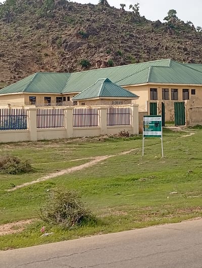 The image depicts a building complex with green roofs, situated against a backdrop of a rocky hillside in Shuwa, Nigeria. The building appears to be a single-story structure with yellow walls and dark-framed windows. A fence, comprised of beige concrete pillars and black metal bars, surrounds the property, with barbed wire along the top. In the foreground, a green sign with white text stands on metal poles, bearing the words "BADAGALI PORTAL EMPOWERMENT HUB." The ground surrounding the building is covered in green grass, with a dirt path winding through it. A paved road runs along the bottom of the image. The environment is dry and rocky, with scattered trees on the hillside. There are no people visible in the image.