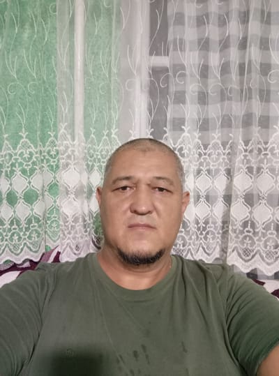 A man with a shaved head and a goatee is pictured from the chest up, facing the camera. He is wearing a dark green t-shirt. He is seated in front of a window covered by sheer, white lace curtains with a green patterned under-curtain. The background suggests an indoor setting, potentially a home in Samat, Kyrgyzstan. The man's expression is neutral, and he is looking directly at the camera.