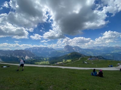 The image captures a scenic view from a grassy hillside in Santa Cristina Gherdëina, Italy, with a vast mountain range in the background under a dramatic sky filled with white and gray clouds. In the foreground, a person wearing a hat, a light-colored shirt, and dark shorts stands on the grass, facing away from the camera and looking towards the mountains. To the left, two individuals are lying on the grass. Further back, several people are seated on the lawn, facing a building complex nestled on a lower slope. A dirt path winds through the grassy terrain, leading towards the buildings. The landscape is characterized by rolling green hills, scattered trees, and the imposing peaks of the Dolomites stretching into the distance.