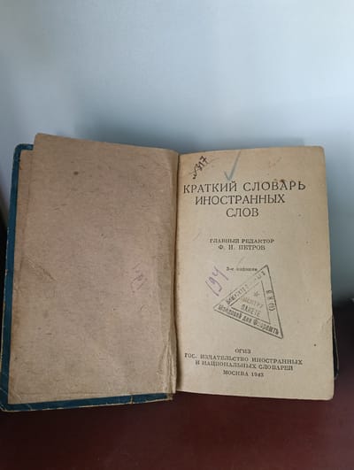 This is an image of an open book, with the right page clearly visible and the left page partially visible. The book appears old and worn, with a faded tan cover. The title of the book, printed in Russian, is "КРАТКИЙ СЛОВАРЬ ИНОСТРАННЫХ СЛОВ," which translates to "Concise Dictionary of Foreign Words." Below the title, it states "ГЛАВНЫЙ РЕДАКТОР Ф. Н. ПЕТРОВ," meaning "Editor-in-Chief F. N. Petrov." Further down, the text reads "3-е издание," indicating it is the "3rd edition." There is a triangular stamp on the right page with Cyrillic text, which appears to be related to a mail or delivery service, possibly mentioning "Центру," meaning "Center," and "Молдова," referencing Moldova. Below the stamp, it states "ОГИЗ ГОС. ИЗДАТЕЛЬСТВО ИНОСТРАННЫХ И НАЦИОНАЛЬНЫХ СЛОВАРЕЙ" which translates to "OGIZ State Publishing House of Foreign and National Dictionaries." The publication year is "МОСКВА 1943" (Moscow 1943). There are handwritten markings on both pages, including "№317" and "194" on the right page. The book is lying on a dark surface, likely a table. The background is a plain, light blue wall. The image does not provide any direct information about the location being the city of Florești, Moldova, other than the potential reference to "Moldova" on the stamp.