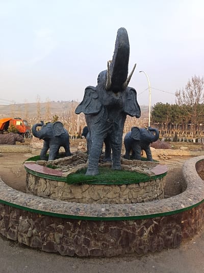 In a public green space within the city of Andijan, Uzbekistan, a central display features three dark-colored elephant statues arranged on a tiered, circular stone structure. A large elephant, positioned centrally, stands with its trunk raised high and tusks prominent. Flanking this larger statue are two smaller elephant statues. The tiered base incorporates artificial green turf and appears to be a dry fountain or decorative planter, surrounded by exposed earth. A curved, low stone wall, constructed from pebbles and irregular stones, defines the immediate foreground. In the mid-ground to the left, a reddish-orange industrial vehicle, possibly a train car or large truck, is visible, along with a partially obscured statue of a brown bear. The background to the left consists of barren deciduous trees and distant hills under a pale, overcast sky. To the right, a mix of evergreen trees and bare deciduous trees are present, alongside a tall, curved street lamp.