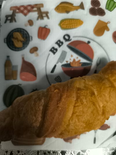 A close-up, overhead shot shows a golden-brown croissant lying on a white napkin with a red and white checkered pattern. The napkin is decorated with various illustrations related to barbecuing, including a grill with flames, a fish, corn on the cob, hamburgers, a bell pepper, chicken pieces on a grill, a ham, a bottle and glass of beverage, mushrooms, and an oven mitt. The background is blurred, but appears to be indoors. The image appears to have been taken in Dillingen an der Donau, Germany.