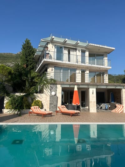 A person is standing on a balcony of a modern multi-story villa in Brca, Montenegro, holding a phone and looking at it. The villa is built with light-colored stone and has large windows and glass railings on its balconies. In front of the villa, there is a swimming pool with clear turquoise water, its surface reflecting the building and the surrounding greenery. On the wooden deck beside the pool, there are two orange and white striped lounge chairs, and an orange umbrella stands partially open near a seating area. The villa is nestled on a hillside with lush green trees and vegetation, with a mountain visible in the background under a clear blue sky.