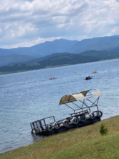 The image depicts a serene lake scene under a bright sky with scattered clouds. In the foreground, a metal structure with a black frame and white canvas covers, possibly a boat rack or a canopy, stands near the water's edge. The structure is adorned with several tires, suggesting a rustic or repurposed design. The lake itself is calm, with gentle ripples reflecting the sky.
Further out on the water, three kayaks can be seen. One kayak, with two occupants, is closer to the shore, while another, with a single occupant, is further away. A third kayak, also with two people, is positioned in the middle distance. The kayakers appear to be enjoying a leisurely paddle on the lake.
In the background, rolling hills covered in dense green foliage rise up, creating a picturesque backdrop. The far distance is characterized by hazy blue mountains, adding depth to the landscape. The overall impression is one of tranquility and outdoor recreation. The time of day appears to be daytime, with clear visibility and bright lighting, suggesting a sunny day. The location cues, such as the type of vegetation and the mountainous terrain, align with the provided context of Tsigov chark, Bulgaria. No visible text is present in the image.