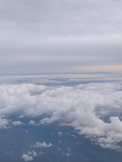 The image shows an aerial view of clouds, possibly taken from an airplane. Thick, puffy white clouds fill the majority of the frame. A layer of lighter gray clouds stretches across the upper portion of the image. In the distance, there is a break in the cloud cover revealing a faint blue area with snow-capped mountains along the horizon. There are no people or discernible objects within the scene. The location in the city of Warngau, Germany is not apparent within the frame.