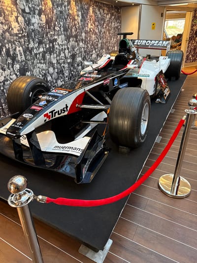 The image shows a Formula 1 race car displayed on a black platform. The car is predominantly black, white, and red, with various sponsor logos visible, including "Trust," "Superfund," and "European." The background features a large black and white mural or photograph of a crowd of people. To the right of the race car, a doorway leads to another area where two men are seated. Red velvet ropes on stanchions are arranged to cordon off the race car display. The floor is made of dark wood planks. The overall impression is that of a museum exhibit or a special display. The image does not provide any specific information that would allow for its localization within the city of Heraklion Municipal Unit, Greece.