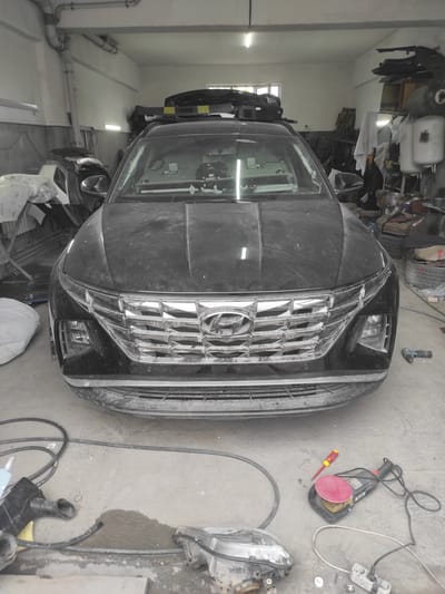 A black Hyundai Tucson is parked in a garage. The car is dusty and appears to be undergoing some kind of repair or detailing. Various car parts, tools, and cleaning supplies are scattered around the vehicle and on the floor. Among the items visible are a car headlight, a sander, a screwdriver, hoses, and what appears to be a compressor or air tank in the background. The garage environment is industrial, with visible pipes and lighting fixtures on the ceiling, and storage units filled with items. The floor is concrete and shows signs of recent activity, with some water or liquid spilled in one area. The overall impression is of a working auto repair shop or detailing bay. The image was taken in the city of Qairat, Kazakhstan.