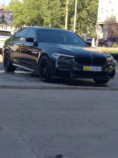 A black BMW sedan is parked on a paved surface in front of a building with trees and a billboard in the background. The license plate of the car reads "07 M2750 R". The vehicle is positioned at an angle, showing its front and side. The environment appears to be an urban setting, likely Yovon, Tajikistan, given the context provided. The car is the central focus of the image.