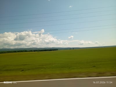 The image depicts a vast, rolling green field under a bright blue sky with scattered white clouds. A line of trees forms a distant horizon, beyond which rolling hills are visible. Several power lines stretch horizontally across the sky. In the foreground, a white stripe marks the edge of a road. The image is a landscape view from a moving vehicle, as evidenced by the slight motion blur. The bottom left corner displays the text "REDMI 13C", and the bottom right corner shows the date and time "16.07.2026 11:34". The location is identified as the city of Galbai, Russia.