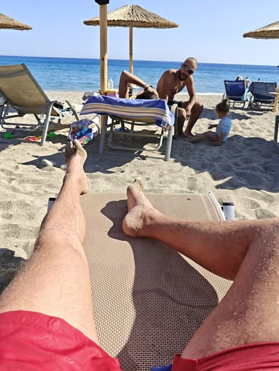 The image captures a sunny day at Plakias beach in Greece. The foreground is dominated by the legs of a person lying on a brown sun lounger. The person is wearing red swim trunks and has sand on their legs and feet.

In the middle ground, a bald man with sunglasses and a beard is sitting on a lounge chair, looking directly at the camera. A child is sitting on the sand near him. Other beachgoers and lounge chairs are visible in the background.

The environment includes a sandy beach, the turquoise sea, and beach umbrellas providing shade. The sky is clear and blue.