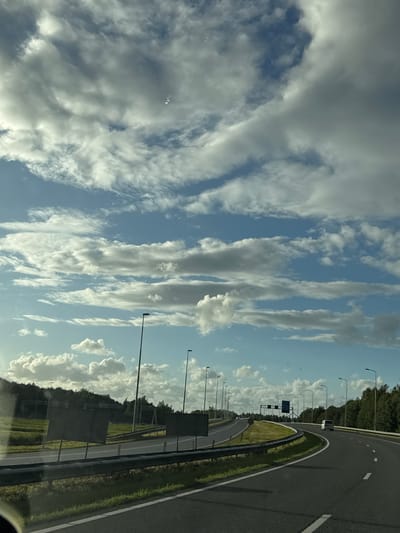 The image shows a view from a vehicle traveling on a highway in Staphorst, Netherlands. The highway has multiple lanes and is bordered by grassy verges and guardrails. There are streetlights lining the road. In the background, there are trees and some structures, possibly road signs or gantries. The sky is partly cloudy with a mix of white and grey clouds. A white van is visible on the highway in the distance. The overall scene depicts a typical highway environment in a rural or semi-rural area.