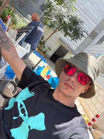 In the city of Tirat Karmel, Israel, a selfie-style shot captures a man wearing a black t-shirt with a large turquoise graphic, a green bucket hat, and red-tinted sunglasses. His left arm, adorned with extensive black tattoos, is visible in the foreground. Behind him, another man with a shaved head wears a navy blue t-shirt with a white logo. They appear to be in an outdoor area with a paved ground surface, possibly a courtyard or street. In the background, there are trees, a building with a tiled facade, and various objects including blue tarps, a white plastic chair, and a red and white striped cone. The sunlight suggests it is daytime.
