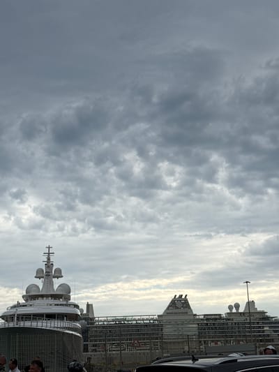 The image shows a partial view of the city of Civitavecchia, Italy, under a cloudy sky. The primary focus is a large cruise ship or yacht docked in the harbor. The ship has multiple levels and a prominent radar or communications mast with several dome-shaped structures. Adjacent to the ship, there is a waterfront area with fences and some infrastructure, and part of a car is visible. The image also captures the surrounding buildings and structures of the city, including a tower with "SILVERSEA" written vertically on it. There are people visible standing in front of the ship, indicating activity on the docks.