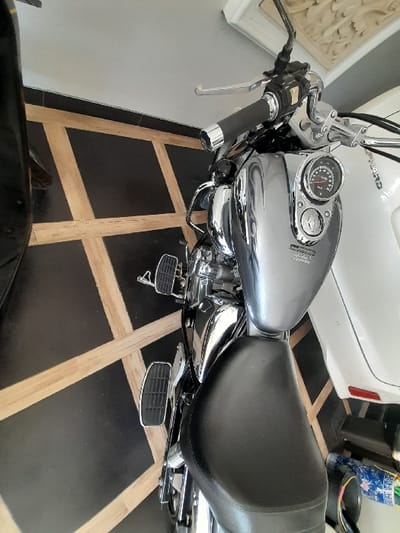A silver-grey Honda cruiser motorcycle, featuring prominent chrome accents on its engine, exhaust, and handlebars, is stationary in what appears to be an indoor or semi-enclosed space. The motorcycle has a black seat and integrated floorboards. Its dashboard, visible on the fuel tank console, includes a speedometer and other gauges. The vehicle is positioned on a black tiled floor with light brown geometric patterns. To the right of the motorcycle, a portion of a white vehicle, likely a car, with a red rear reflector is visible. A section of a black vehicle is discernible on the far left. The background consists of a light-colored wall, with an ornate, white carved architectural detail positioned in the upper right. No individuals are present in the scene. This scene is located in Entalsewu, Indonesia.
