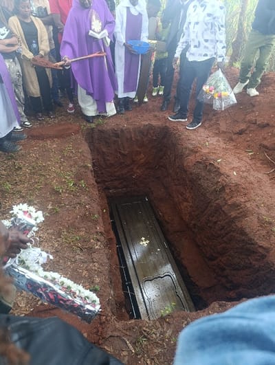 A burial is depicted outdoors in Muthetheni ward, Kenya. A dark-colored casket with a golden cross emblem rests within a rectangular grave dug into reddish-brown soil, partially submerged in water. Approximately 10-15 individuals are gathered around the grave. A clergyman in a purple chasuble and white alb holds an open book and a wooden stick with soil, extending it over the grave. Another clergyman in a white alb holds a blue bowl. Attendees include a woman in a light brown cardigan and a man in a white patterned shirt holding a bouquet of multi-colored flowers wrapped in clear plastic. In the foreground, hands hold a bouquet of red and white flowers wrapped in silver foil. The ground is moist, and green vegetation is visible in the background.