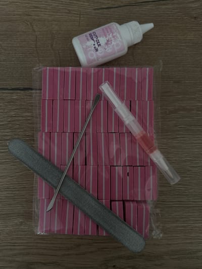 The image displays a collection of nail care essentials laid out on a light brown wooden surface, showing distinct wood grain patterns.
The main subjects are various manicure tools and products. On the left, there is a white plastic bottle with a conical white spout. Its label is light purple/pink with white text, partially readable as "Lavender," "Aloe," "Moni..." (likely part of a brand or ingredient list), "CUTICLE REMOVER," and "40 ml."
To the right of the bottle, centered in the upper portion of the frame, is a clear plastic pen-like applicator containing a reddish-pink liquid, presumably cuticle oil, with a brush tip visible at one end.
Dominating the central and right-hand side of the image is a large stack of small, rectangular nail buffers. These buffers are pink with thin white horizontal stripes and appear to be packaged together in clear plastic.
Overlapping the stacked buffers are two metal tools. A dark grey nail file with a coarse texture and a rounded end is positioned diagonally across the lower right. Lying on top of the buffers and crossing the nail file is a thin, metallic, double-ended cuticle pusher, with one end shaped like a small spoon and the other a slightly curved blade or pusher.
No people are present in the image, and no specific activity is taking place; it is a static flat lay arrangement of items. The setting appears to be indoors, given the uniform lighting and wooden surface. The time of day or weather is not discernible. The visible text confirms the presence of a "CUTICLE REMOVER" in a "40 ml" bottle.