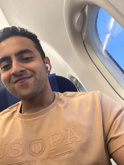 A man sits in an airplane seat with his head tilted, smiling at the camera. He has dark hair and is wearing a light brown t-shirt. A white earbud is visible in his ear. The airplane window with bright blue sky is in the upper left. The seat is navy with blue trim. The ceiling of the plane is white.