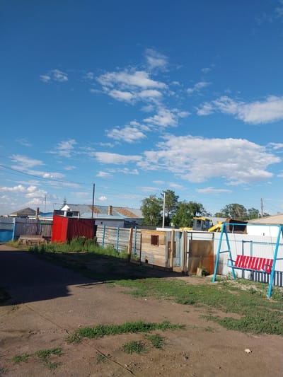 This media file depicts an outdoor scene in Трудовое, Kazakhstan, under a clear blue sky with scattered white clouds. The image is oriented vertically, giving a sense of looking upwards and sideways.
The foreground is dominated by a dusty, unpaved ground with patches of green grass. A dark shadow, likely cast by a structure or object, stretches across the ground. A thin, rusty wire runs diagonally across the dirt.
In the mid-ground, there are several rustic, makeshift structures that appear to be houses or sheds, constructed with corrugated metal and wooden planks. One prominent structure is painted red, contrasting with the neutral tones of the others. A blue and red swing set is visible in the upper left portion of this area. Behind the structures, trees with green foliage are present, suggesting a rural or semi-rural environment. A yellow excavator is partially visible behind some of the buildings.
The background is a vast expanse of blue sky filled with fluffy white clouds. The lighting suggests it is daytime, likely late morning or afternoon, given the shadows. There are no people visible in the image. The overall impression is one of a simple, perhaps developing, community or a backyard area.
