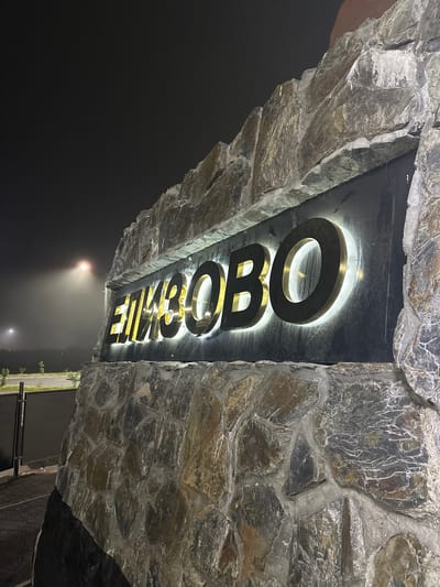 This media file shows a sign with illuminated gold lettering against a dark background, set within a rustic stone structure. The sign is oriented vertically and appears to read "КРАСНОЕ" (Krasnoe), which is a place name. The stone structure is composed of irregular, dark gray and brown stones with visible mortar between them. The letters of the sign are three-dimensional and have a metallic sheen, with a bright white light emanating from behind them, creating a halo effect. The image is taken at night, as indicated by the darkness and the presence of artificial lighting. In the background, a soft, diffused light suggests fog or mist, with a streetlamp visible in the distance. The overall atmosphere is somewhat mysterious and perhaps a bit dramatic due to the low light and the rugged texture of the stone. There are no people visible in the image. The setting appears to be an entrance or a landmark.