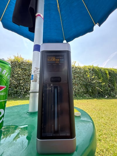 This media file captures an outdoor scene on a sunny day, likely in Vicenza, Italy. The main focus is a modern electronic device, a CUKTECH power bank, positioned on a green plastic table. The power bank has a digital display showing "95%" battery, indicating it's almost fully charged. Text on the device also mentions "OUTPUT", "Type-C1", "Type-C2", and "USB-A", suggesting its charging capabilities.
Adjacent to the table, a blue umbrella provides shade, with its fabric and yellow spokes visible. A silver pole, part of the umbrella or another structure, runs through the frame and has decorative purple and pink tape wrapped around it. A black piece of fabric, possibly clothing, hangs from the pole. In the background, a lush green hedge and a well-maintained lawn suggest a garden or park setting. The sky is a clear blue with faint white clouds.
Adding a touch of casual leisure, a green can of Heineken beer is visible in the foreground, partially obscuring the table and the hedge. The overall impression is a relaxed outdoor moment, possibly a break during a warm afternoon. The bright sunlight and clear sky point to fair weather. The text on the beer can includes "ORIGINAL" and "AMSTERDAM 1873", further confirming its brand.
