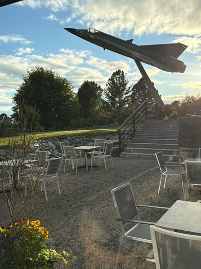 The image shows an outdoor seating area with gravel flooring, furnished with several metal tables and chairs. In the background, a set of outdoor stairs with a dark railing leads upwards. Dominating the upper portion of the scene is a static display of a military jet fighter, mounted on a stand, angled upwards against a cloudy blue sky. To the left of the stairs and jet, there are trees and a grassy slope. In the foreground, to the left, a planter contains colorful flowers. The overall setting appears to be a publicly accessible area, possibly a restaurant or park, given the outdoor dining setup and the prominent aircraft display. This scene is located at the Östersund Aircraft Museum (Flygvapenmuseum Östersund) in Östersund, Sweden, which features static displays of aircraft.