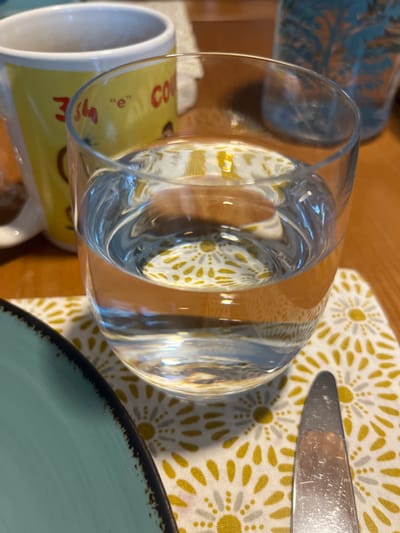 This close-up image, rotated 90 degrees clockwise, presents a detailed view of a dining table setting. In the foreground, a clear, empty glass lies horizontally across the frame. Its cylindrical form distorts the underlying white placemat, which features a repeating pattern of yellow/gold and gray floral or starburst designs.
To the right of the glass, a silver-colored knife with a smooth blade is partially visible, resting on the placemat. In the bottom right corner, a light blue or turquoise plate with a distinct dark, textured rim is partially seen. The reflections of light are visible on the glass and the knife, suggesting indoor lighting.
In the bottom left, a white ceramic mug with a prominent yellow-gold band is partially visible. Red text is printed on this band, with "CO" and a distinct lowercase "e" clearly discernible, hinting at words like "COFFEE." Behind the mug and glass, an out-of-focus object, possibly another patterned plate or dish with a teal-like color and intricate design, can be seen in the upper left background. The visible table surface beneath the items appears to be light-colored wood. The overall scene suggests a prepared table, possibly for a meal or beverage, in an indoor setting. There are no people visible. The context of Chêne-Bourg, Switzerland, is provided, but no specific visual cues in the image confirm this location.