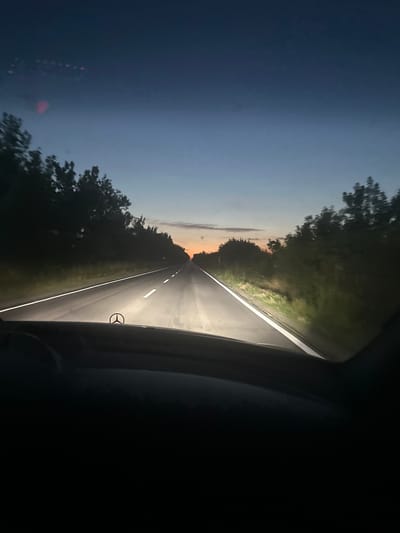 The image shows a view from the perspective of someone driving a car on a road at dusk. The road ahead is illuminated by the car's headlights, with white lane markings visible. Trees line both sides of the road, silhouetted against the twilight sky. The sky transitions from a deep blue at the top to lighter shades of orange and yellow near the horizon, indicating the presence of a sunset or sunrise. A Mercedes-Benz emblem is visible on the hood of the car in the foreground. The scene is set in a rural or suburban area, outside of the city of Manastir, Bulgaria. There are no people visible in the image.