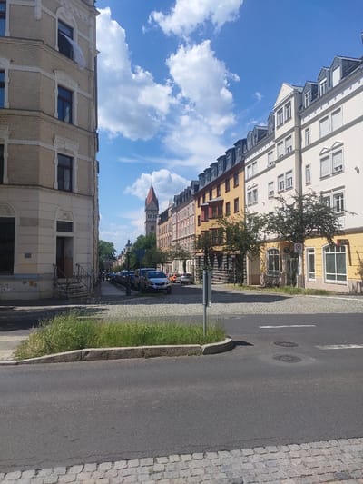 The image shows a street scene in Chemnitz, Germany, on a sunny day with a blue sky and scattered clouds. The street is paved with asphalt in the foreground and cobblestones further back. Several parked cars are visible along the sidewalk. Buildings line the street on both sides, with the left side featuring a light-colored, ornate building and the right side displaying a row of multi-story buildings with pitched roofs and dormer windows. A church tower with a red roof is visible in the distance. The scene appears to be a residential or commercial area.