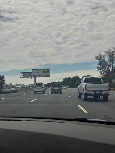 This is a forward-facing, eye-level, medium shot captured from inside a vehicle on a highway in Fairfield, California, USA, on an overcast day. The highway has multiple lanes, and the road surface is asphalt. White lane markings are visible.
In the foreground, the dashboard of the vehicle from which the photo is taken is partially visible. In the middle ground, several vehicles are on the highway. From left to right, a white SUV, a black SUV, and a white pickup truck are present. The white pickup truck is the most prominent vehicle in the frame and is driving in the right lane. Its license plate reads "05629C2".
In the background, there is a large overhead electronic sign displaying "EXPRESS LANE", "TO WEST", "TOLL 0.75", and "OPEN 7 DAYS 5AM - 8PM". There are also trees lining the side of the highway, and in the far distance, mountains are visible under a cloudy sky. The clouds are dense and cover most of the sky, with patches of blue sky peeking through.