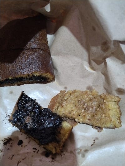 This media file shows two pieces of martabak, a type of stuffed pancake popular in Indonesia. The martabak pieces are placed on brown paper.
The first piece, in the upper left, is larger and appears to be a whole or half martabak cut into a rectangular shape. It has a dark brown, slightly textured outer layer and a thick, dark filling, likely chocolate sprinkles and condensed milk, visible in its cross-section. The second piece, in the lower half of the image, is smaller and triangular, cut from a different martabak. This piece is also dark brown on top, but the exposed interior reveals a lighter, more crumbly texture. To the right of this piece is another section of martabak with a lighter, golden-brown exterior, sprinkled with what appears to be sugar and possibly chopped peanuts or other nuts. The lighting suggests this image was taken indoors, possibly in a home or street food stall, during the day.
The scene is casual, focusing on the food itself. The presentation is simple, with the martabak resting on a common food wrapping material. There are no people or significant environmental details visible to indicate a specific time of day or weather conditions, other than the indoor lighting. No text is visible in the image. The context provided places this scene in Palangka Raya, Indonesia, a region where martabak is a common street food and dessert.